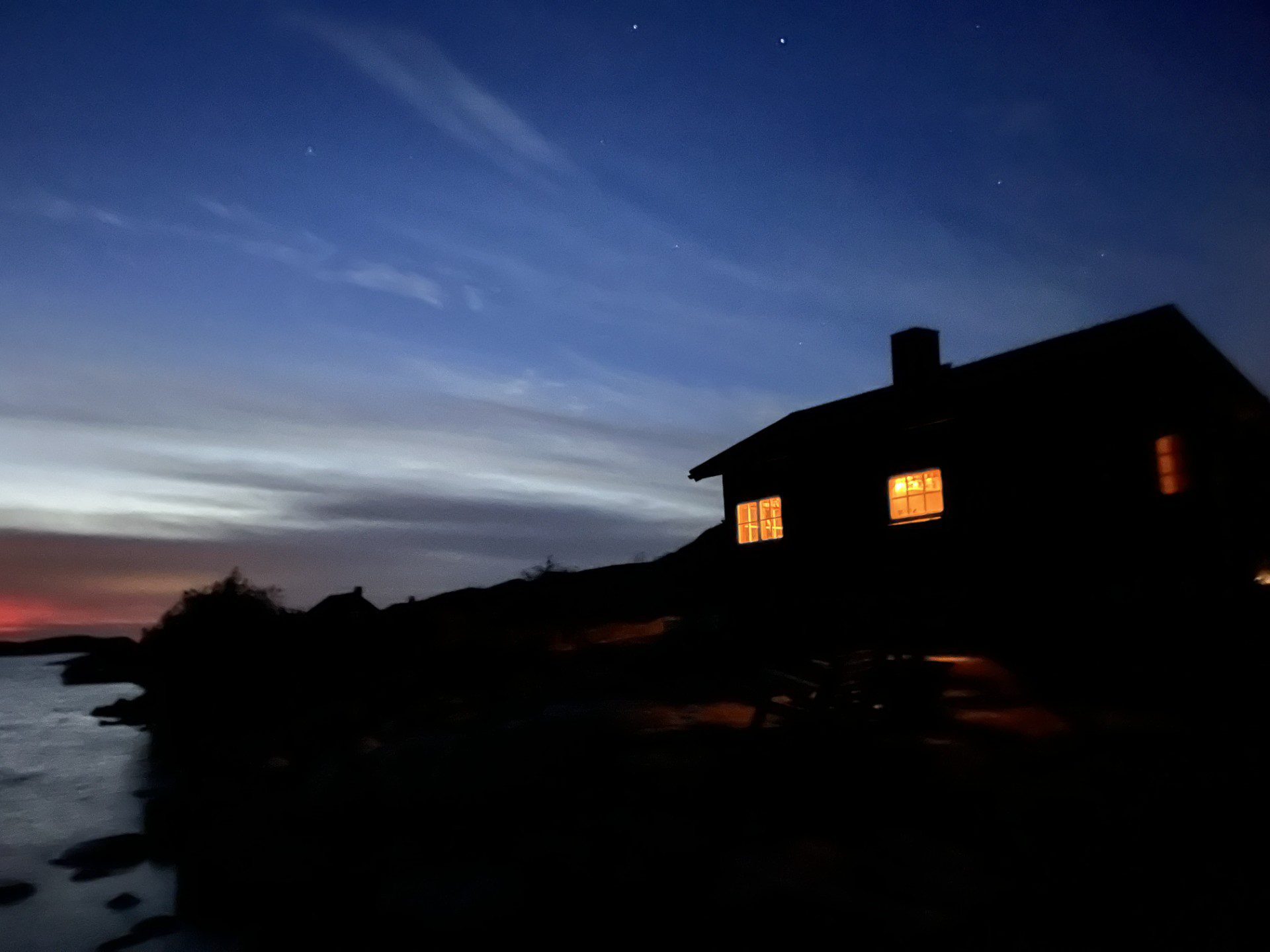 Båttransfers till öar i Stockholms skärgård A traditional fishing and hunting lodge on the island of Fredlarna at night with warm, glowing window lights, seen from the sea during a private RIB charter in the Stockholm archipelago with 59 DEGREES NORTH.